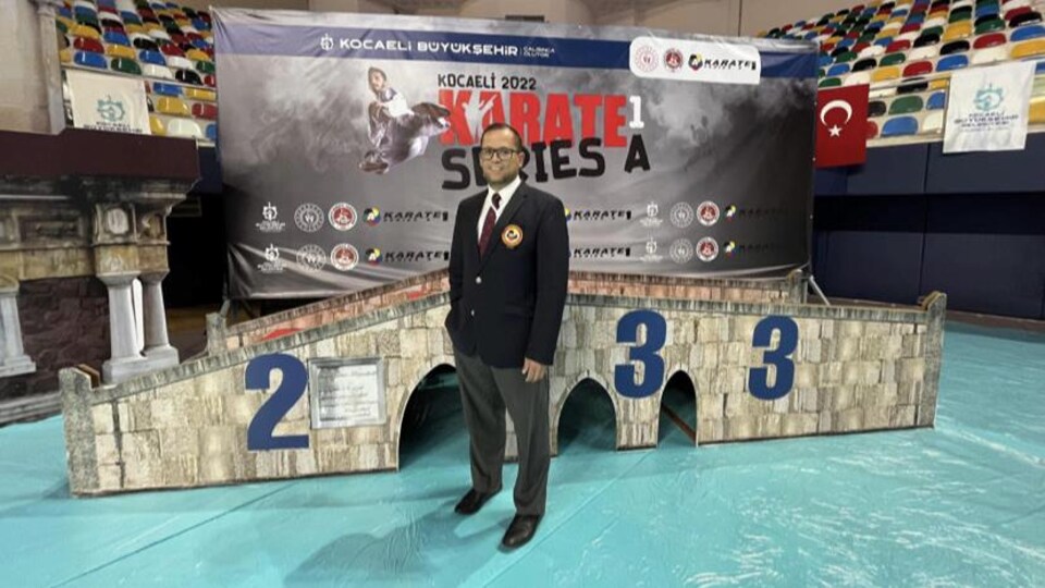 Samir Baccouche, posant dans un amphithéâtre, debout devant l'affiche de la compétition.