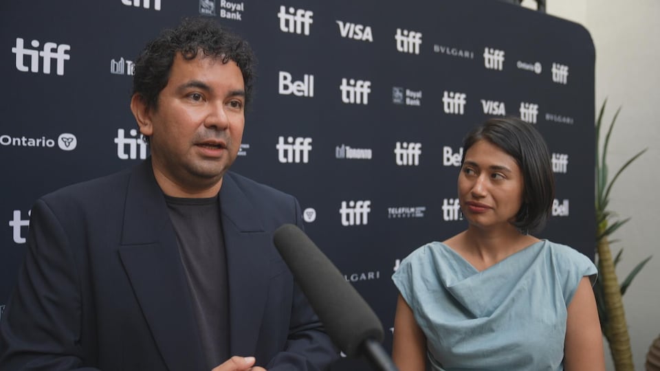 Jason et Kelly, debout devant un panneau avec le logo du TIFF, répondent à des questions en entrevue.