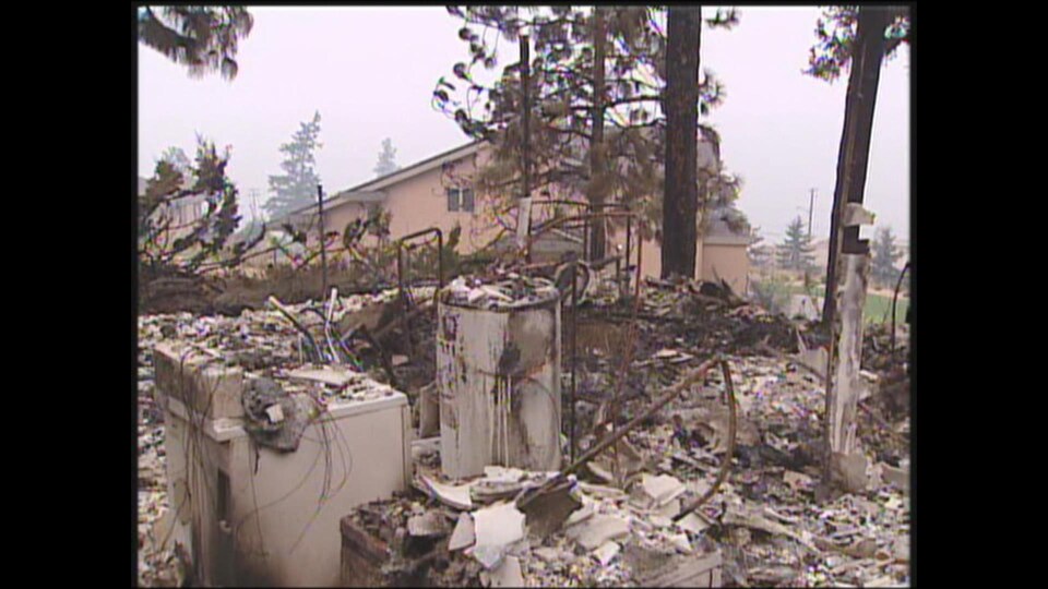 Un poêle et un chauffe-eau endommagés par le feu au milieu d'une pile de débris, avec une maison et quelques arbres à l'arrière-plan.