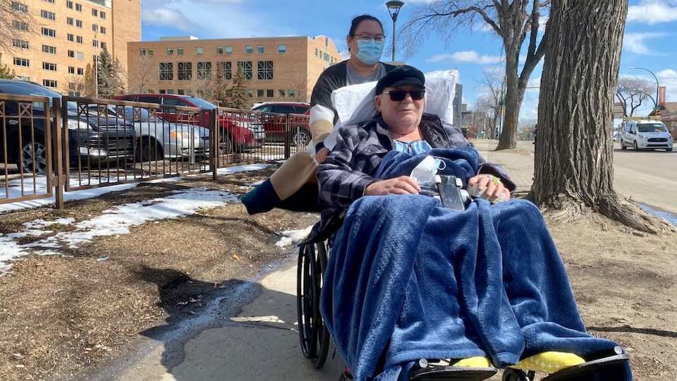 A woman pushed a wheelchair occupied by a man on the sidewalk near a hospital.