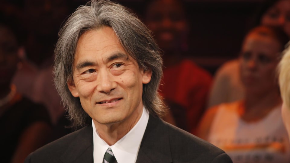 Plan buste de Kent Nagano sur un plateau télévisé. Des gens sont à l'arrière-plan.