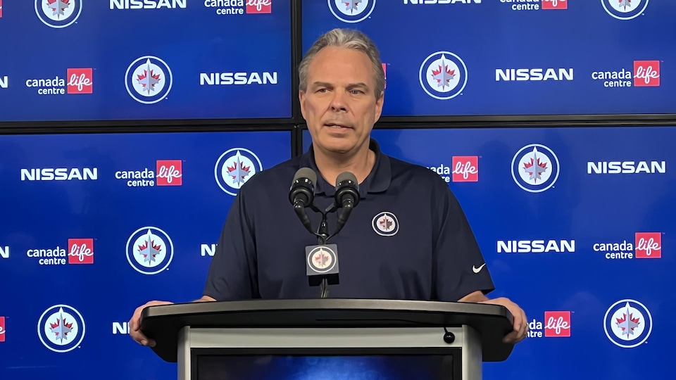 Kevin Cheveldayoff est au podium et parle aux médias. Il porte un polo des Jets de Winnipeg et derrière lui se trouvent des logos de l'équipe et des commanditaires.