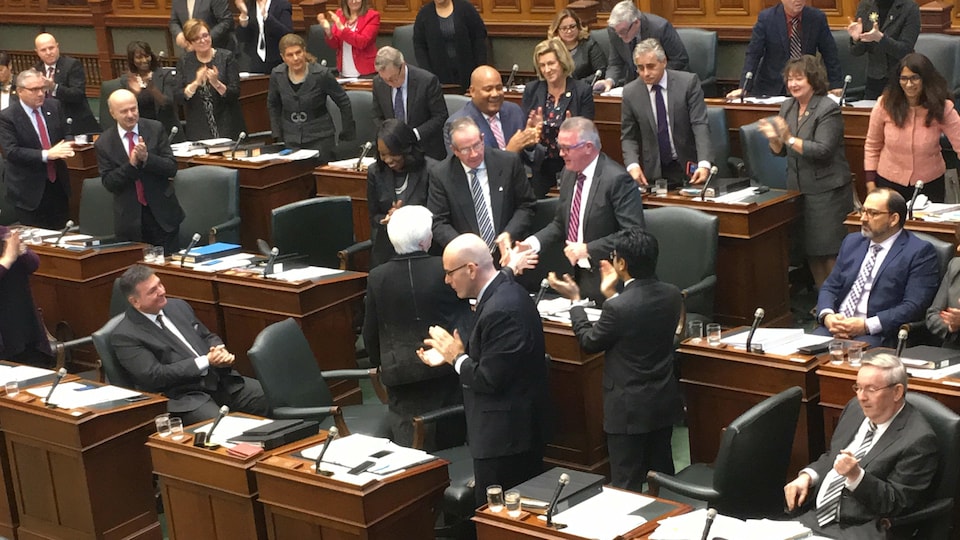 Le ministre est debout en chambre et se fait serrer la main par une député, alors que les autres élus autour applaudissent.