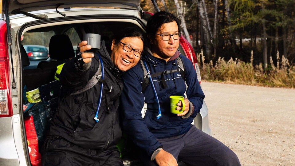Kevin Papatie et Drake sont assis sur la valise ouverte d'une voiture. Kevin Papatie lève son verre en souriant.