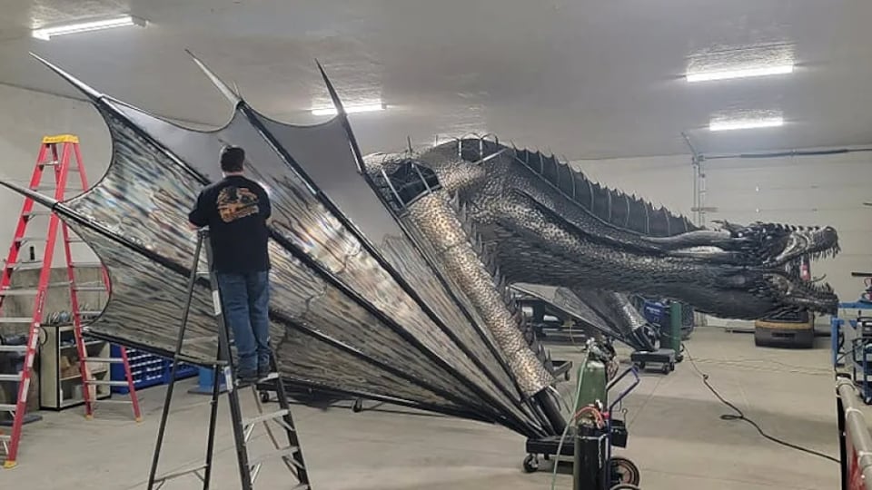 Un homme est installé sur une échelle devant un immense dragon métallique dans un garage.