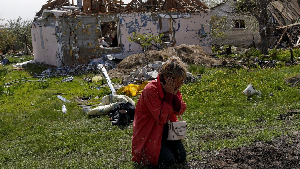 Une femme pleure, à genoux et le visage dans ses mains, devant une maison bombardée.