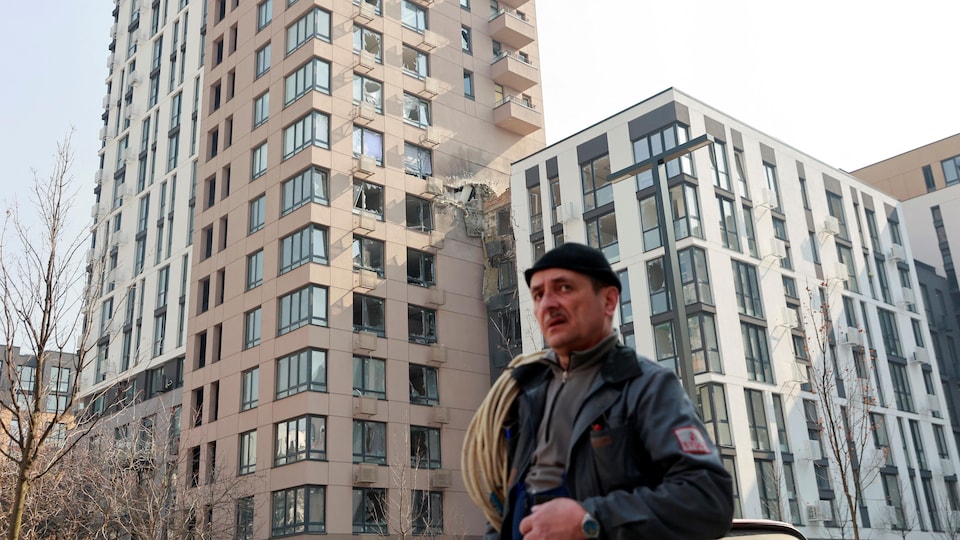 Un homme près d'une tour d'une douzaine d'étages, dont un mur a été percé. Plusieurs fenêtres de l'immeuble ont volé en éclats. 