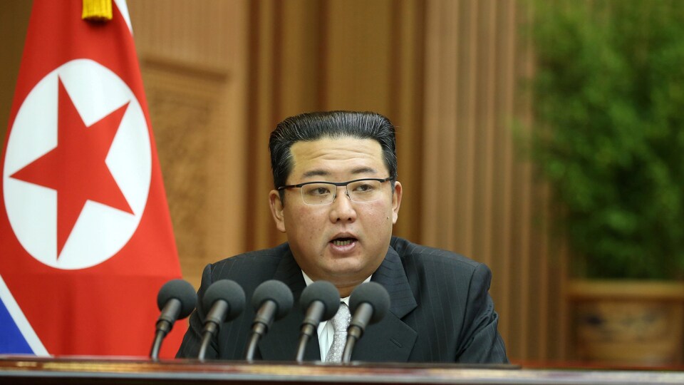 Il parle dans cinq micros devant un drapeau de la Corée du Nord.