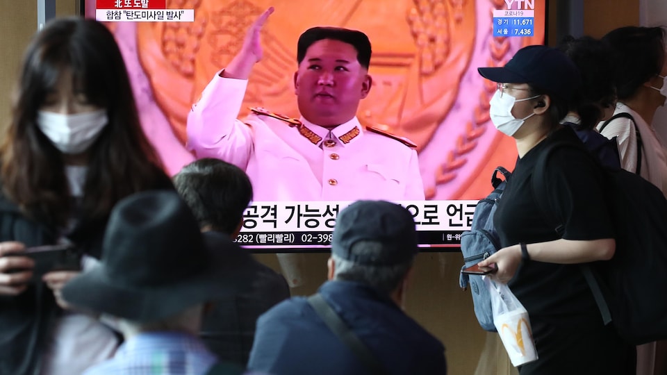 Des personnes portant un masque interagissent devant une télévision qui présente Kim Jong-un. 