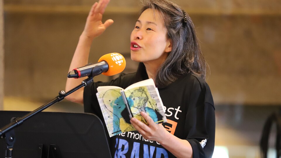 Kim Thúy parle dans un micro avec un carnet à la main. Elle a la main en l'air, signe de l'intensité des émotions. 