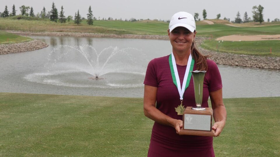 Kim Walker, vêtue d'une casquette sur un terrain de golf, sourit et porte une médaille au cou, tenant son trophée de championne du tournoi de golf.