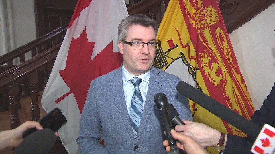 Kris Austin en mêlée de presse devant des drapeaux du Nouveau-Brunswick et du Canada.