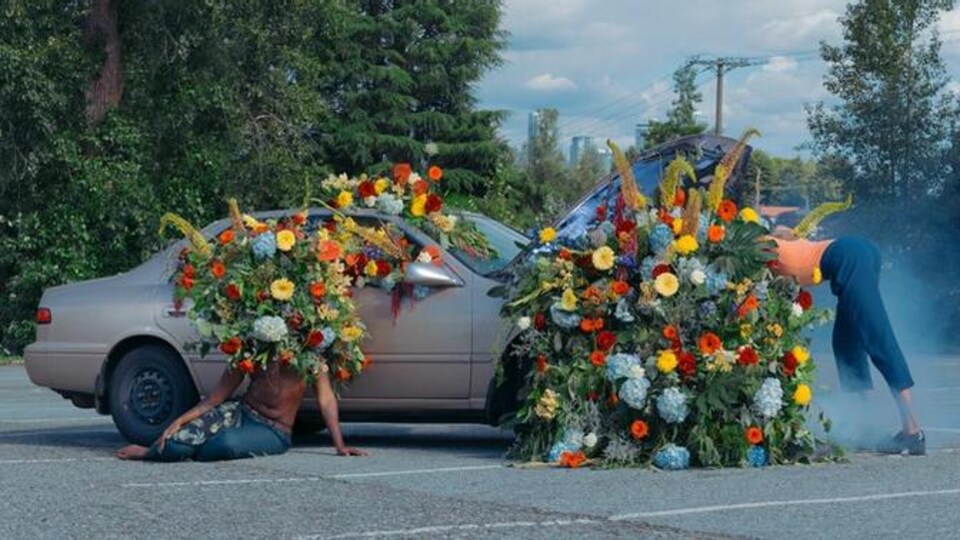 une voiture en panne remplie de fleurs. une personne est assise devant, avec des fleurs à la place de la tête, l'autre est penchée sur le moteur.