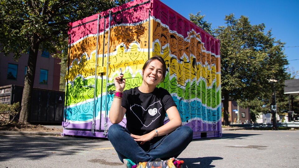 Une femme est assise sur la chaussée devant un conteneur aux couleurs de l'arc-en-ciel.