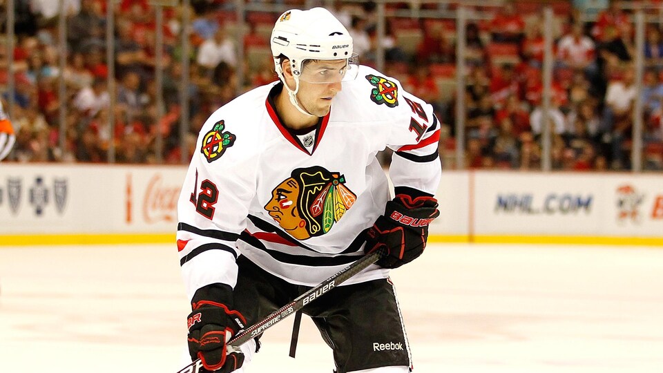 Un joueur de hockey droitier, vêtu de l'uniforme blanc et noir des Blackhawks de Chicago, s'apprête à patiner pendant un match préparatoire de la LNH. 