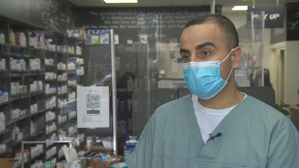 Kyro Maseh répond aux questions, un masque chirurgical sur le visage avec derrière lui sa pharmacie.