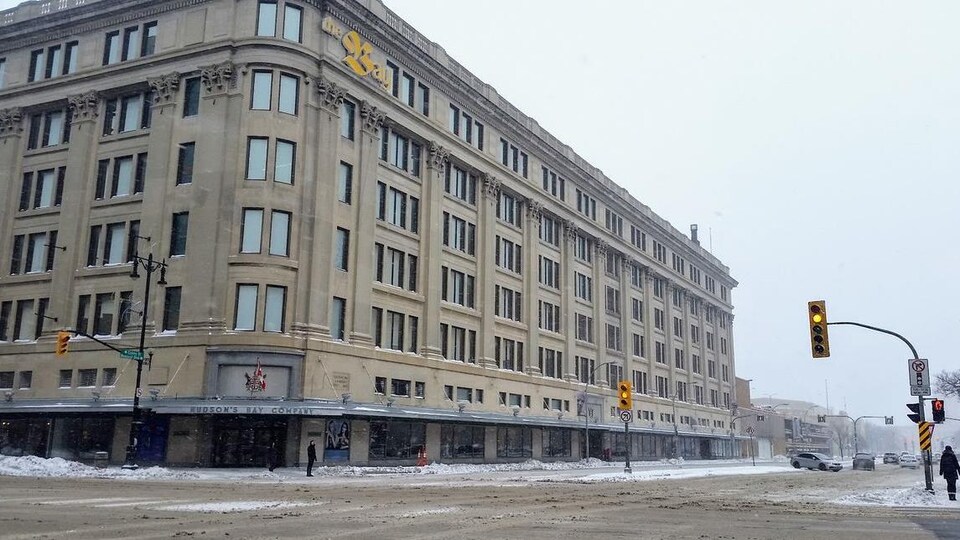 Un grand immeuble brun-gris, vu de l'extérieur, à l'angle de deux rues passantes, un jour de neige.