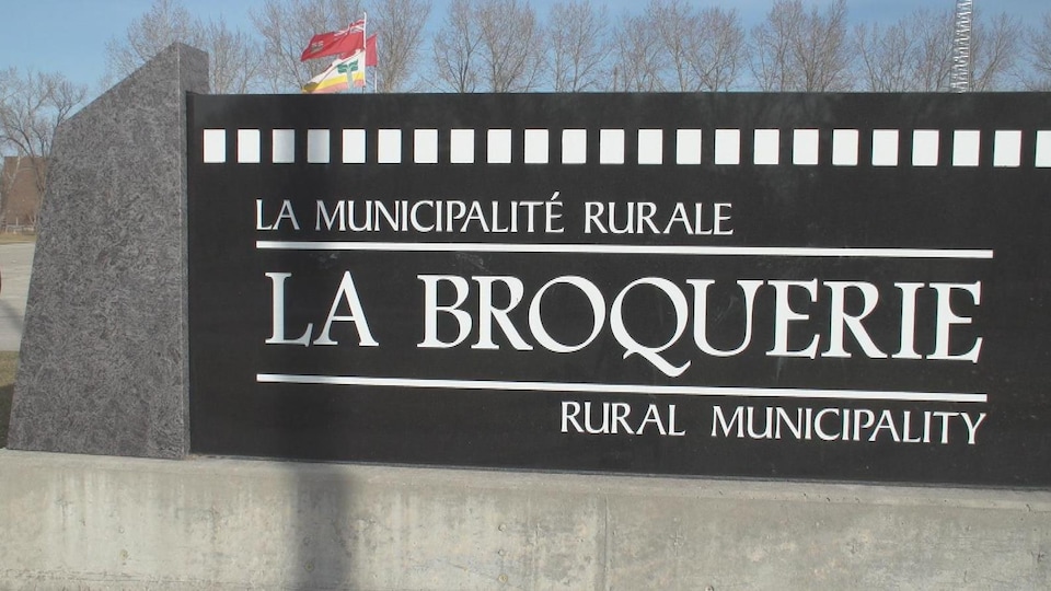 Un homme masqué marchant à côté du panneau de la municipalité rurale de La Broquerie.