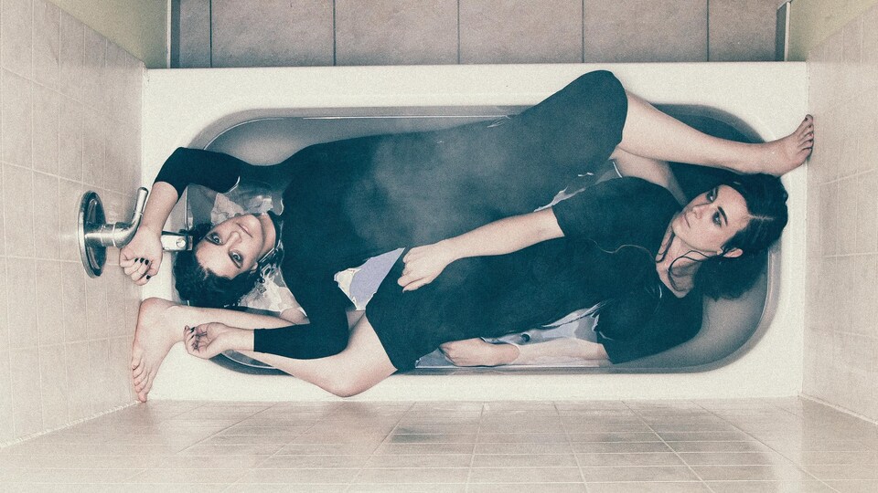 Les deux femmes sont couchées habillées dans un bain et elles regardent la caméra qui se trouve au plafond.