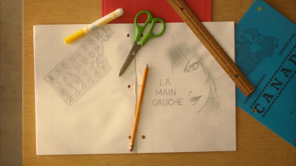 Dans un cahier Canada, un dessin au plomb du visage d'une femme et le titre « La main gauche ». Sur le cahier, un crayon de bois, des ciseaux, un crayon-feutre et une règle en bois.