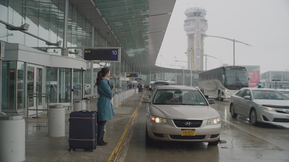 Une femme est debout à l'extérieur d'un aéroport avec une valise à côté d'elle.