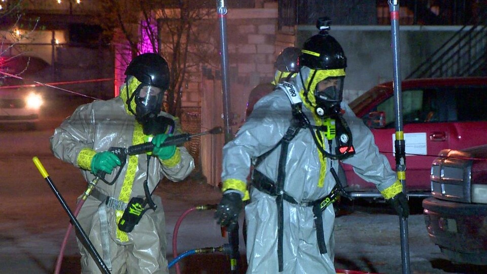 Deux hommes vêtus de chienne, de gants, de masque et d'un équipement pour décontaminer les lieux marchent vers le garage.
