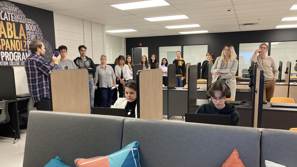 Attablés à des postes de travail numériques individuels et entourés de plusieurs personnes, des jeunes regardent des ordinateurs dans une salle.