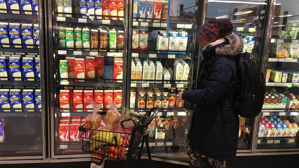 Une femme poussant un petit chariot d'épicerie passe devant des réfrigérateurs contenant du lait dans un supermarché.