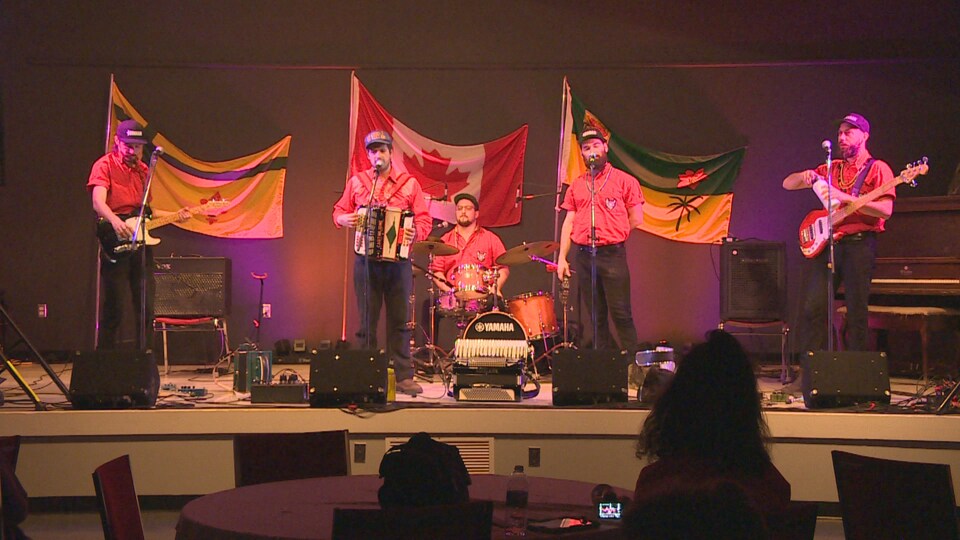 Cinq musiciens sur la scène, devant trois drapeaux, soit le drapeau fransaskois, le drapeau canadien et le drapeau de la province. 