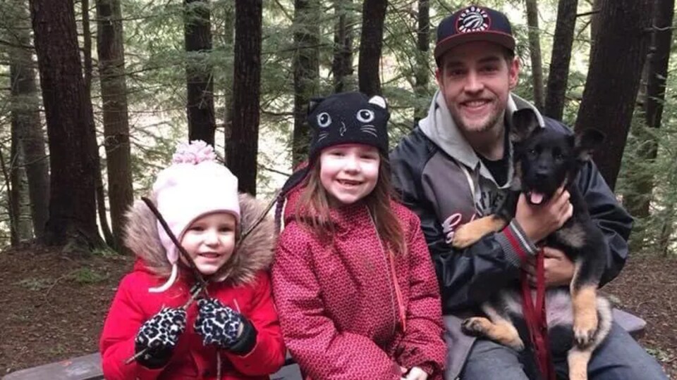 Luke Landry assis sur un banc à côté de ses deux fillettes tient un chien dans ses bras.