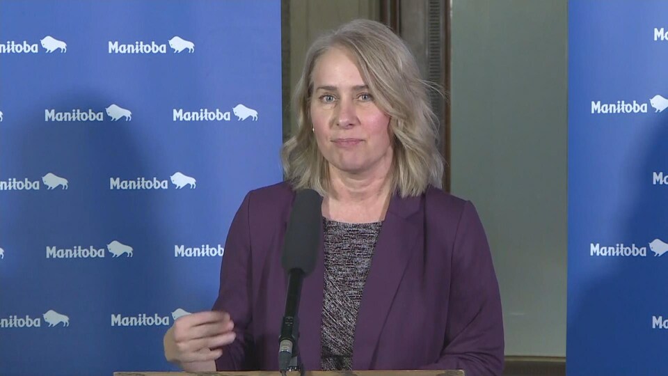L'infirmière en chef de Soins communs Manitoba, Lanette Siragusa, lors d'une conférence de presse devant deux rideaux bleus avec le logo du Manitoba. 

