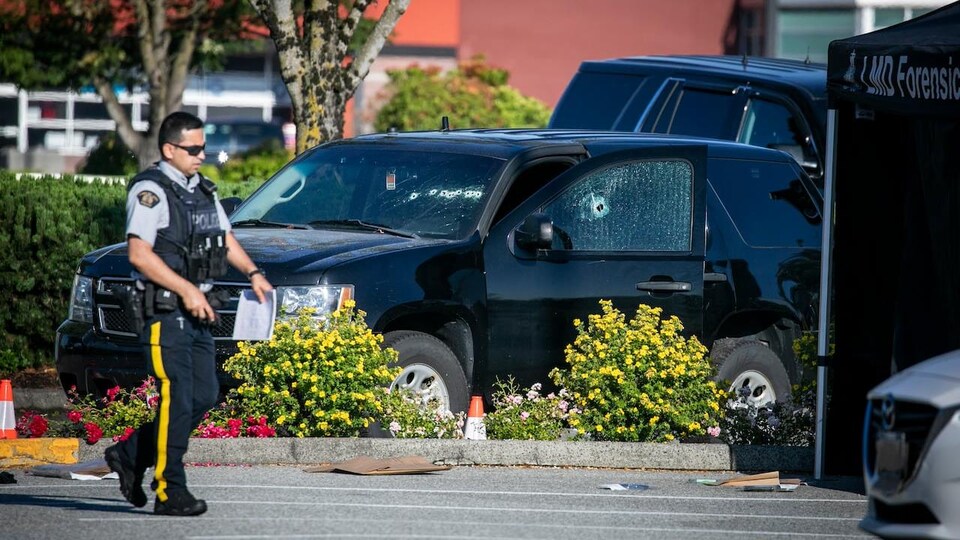 Un policier marche devant une voiture avec un impact de balle. 