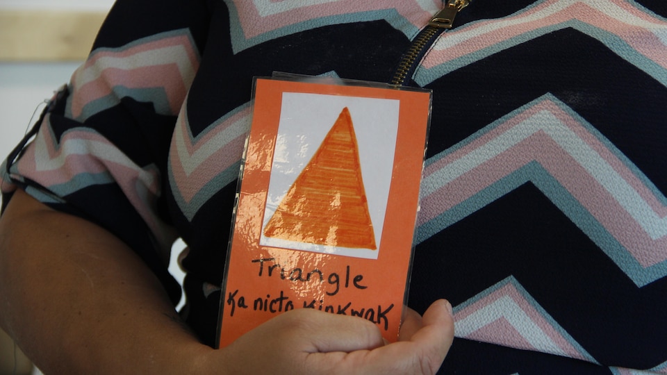 Une femme tient dans la main un papier sur lequel est dessiné un triangle. Y est inscrit le mot "triangle" et l'équivalent en atikamekw. 