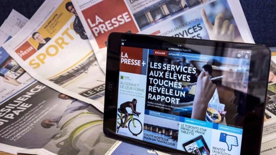 L'application La Presse + est ouverte sur une tablette électronique, devant des cahiers de l'édition papier de La Presse.