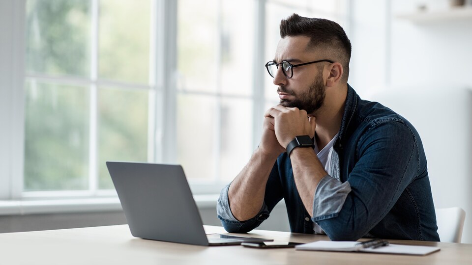 Un jeune homme portant des lunettes regarde l'ordinateur portable posé devant lui. Il est assis à une table ou un bureau, sur lequel sont posés un téléphone cellulaire et un calepin.