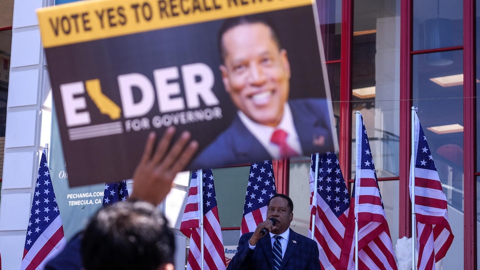 Devant des drapeaux américains, Larry Elder s'adresse à des partisans, qui brandissent une pancarte à son effigie.