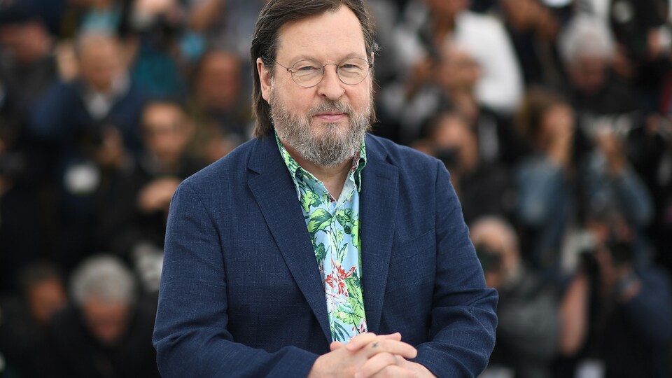 L'homme qui porte des lunettes a les mains croisées sur un pupitre devant lui. Il sourit.