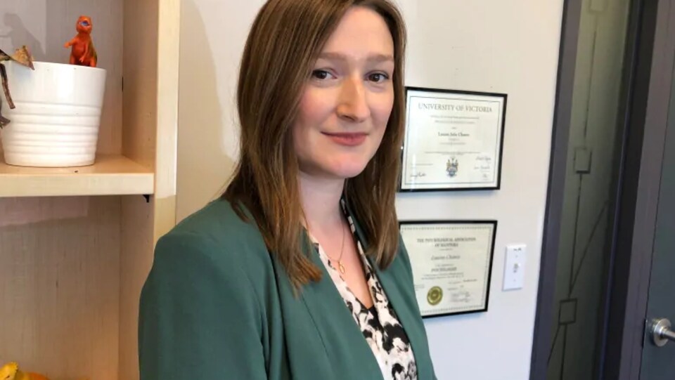 Une femme sourit dans une pièce. Derrière elle se trouve une étagère et des diplômes sont accrochés au mur.