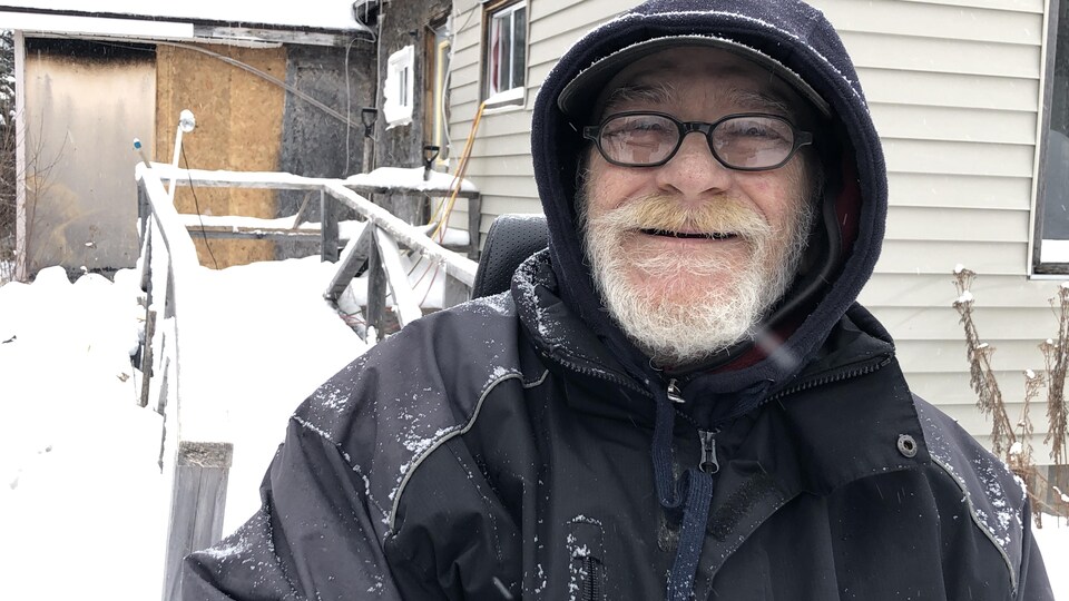 Un homme en manteau sourit à la caméra devant une maison mal en point.