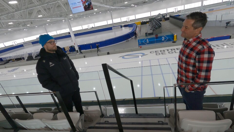 Deux hommes face à face, dans les gradins d'un centre de glace, discutent.