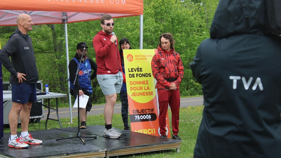 Laurent Dubreuil speaks into the microphone on a small stage, Miha Fontaine and Lu Richer by his side.