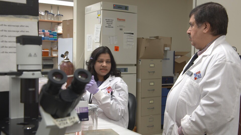 Suresh Tikoo, on the right, who is wearing a lab coat, talks to a researcher, on the left, who is also wearing a lab coat. There is a microscope in the foreground.