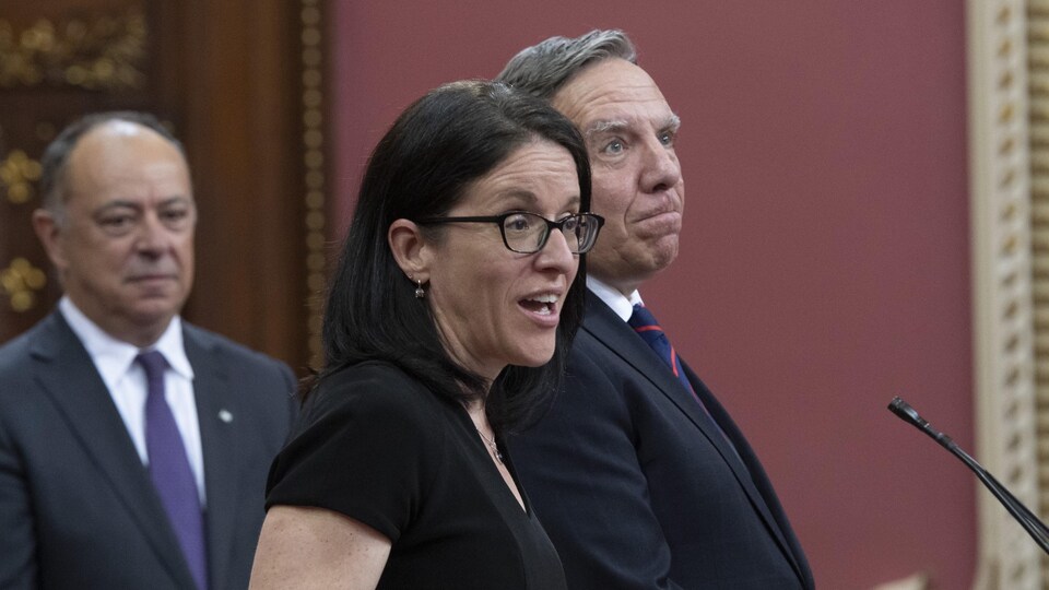 Plan moyen de Sonia LeBel parlant derrière un micro. François Legault et Christian Dubé sont en arrière-plan.
