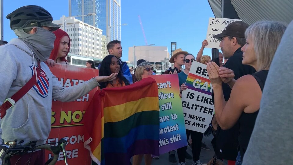 Contre-manifestants portant des affiches et des drapeaux LGBTQ faisant face aux manifestants qui les filment. 
