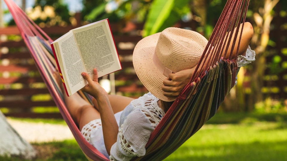 Une femme avec un chapeau lit un livre dans un hamac.