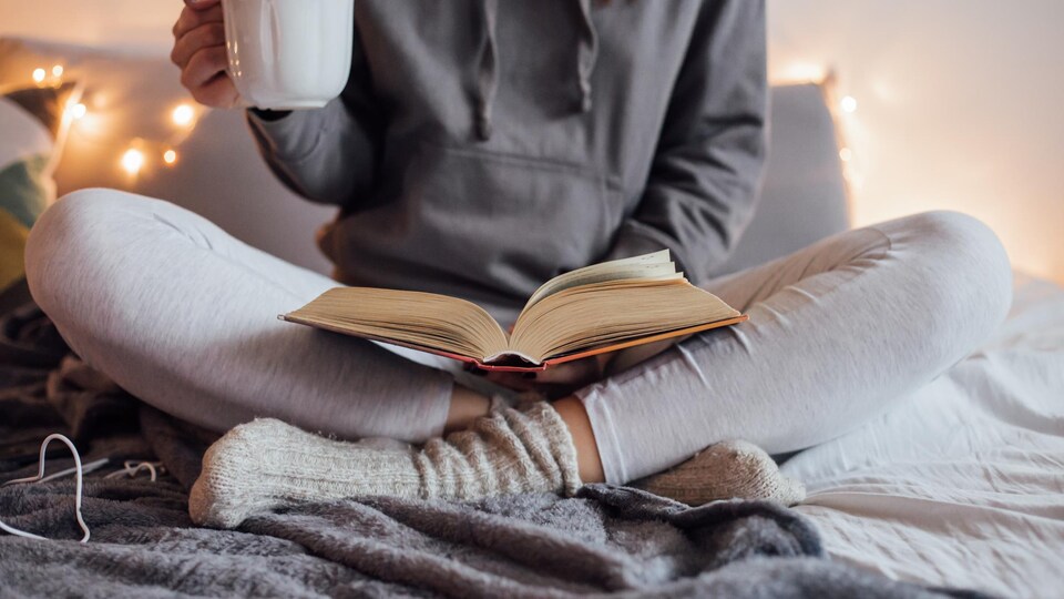 Une femme lit un livre au lit avec une boisson chaude dans une main.