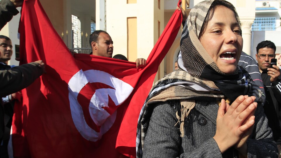 Des manifestants brandissent derrière elle le drapeau national tunisien.