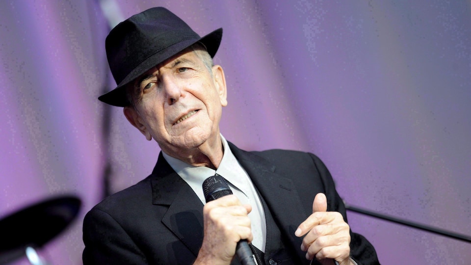 Le chanteur Leonard Cohen portant un chapeau noir et un complet, micro en main, sur une scène extérieure.