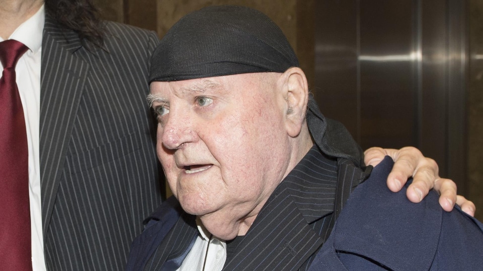 Photo d'un vieil homme portant un manteau bleu et un complet noir et un foulard noir sur la tête.