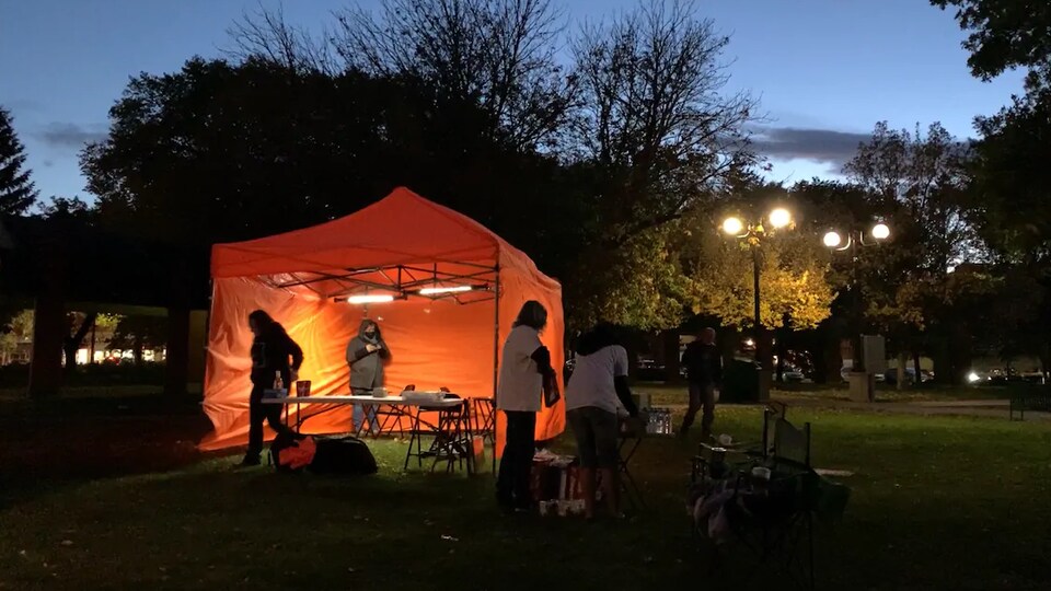 Une tente orange installée au milieu d'un parc avec des bénévoles à l'intérieur, à la tombée de la nuit. 